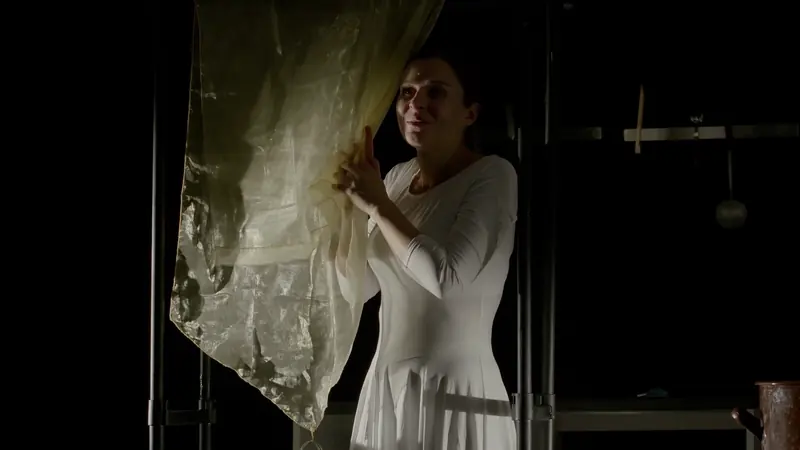 Une femme en robe blanche écarte un rideau de dentelle sur une scène plongée dans le noir.