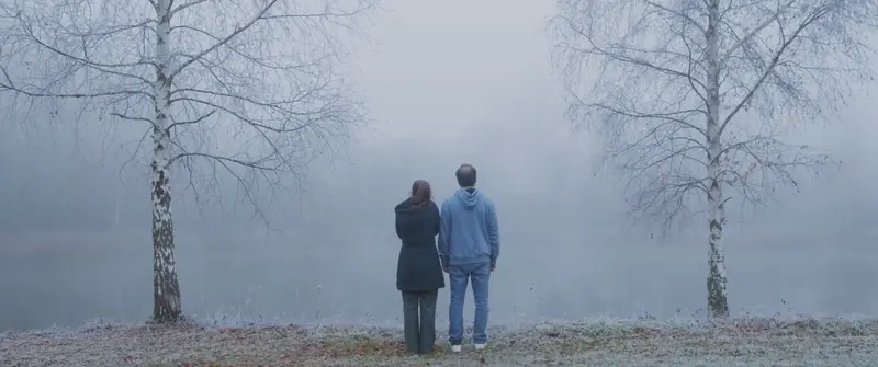 Un couple de dos au bord d’un lac, entre deux bouleaux, dans une brume bleutée.