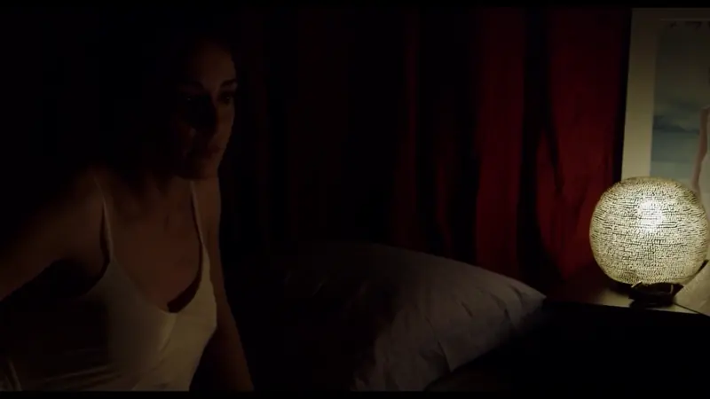 Une femme dans une chambre sombre, éclairée par une lampe globe devant un rideau rouge.