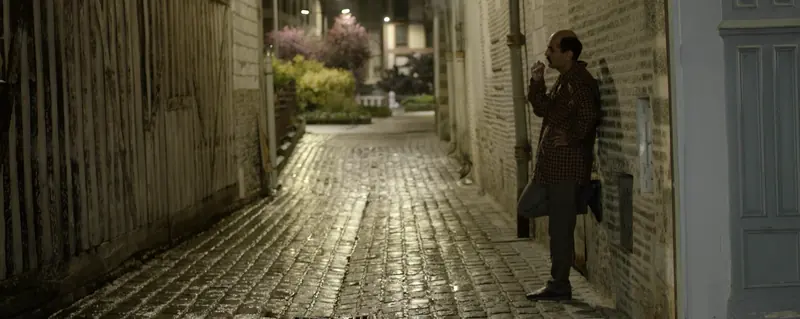 Un homme adossé au mur d’une ruelle pavée, éclairée par des lampadaires, la nuit.